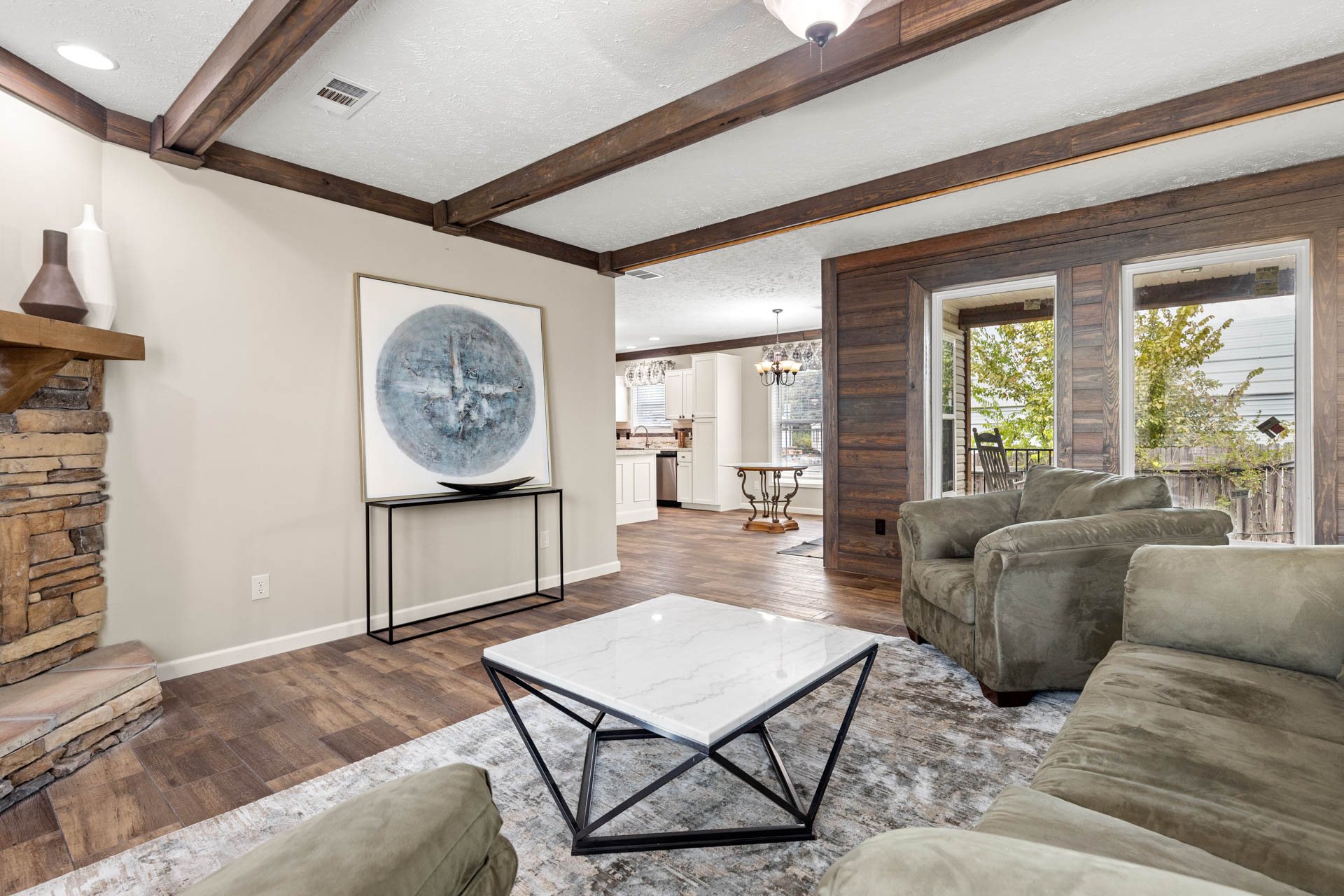 Cozy living room with modern rustic style; features wood-beamed ceiling, stone fireplace, abstract art, suede sofas, and a marble coffee table.