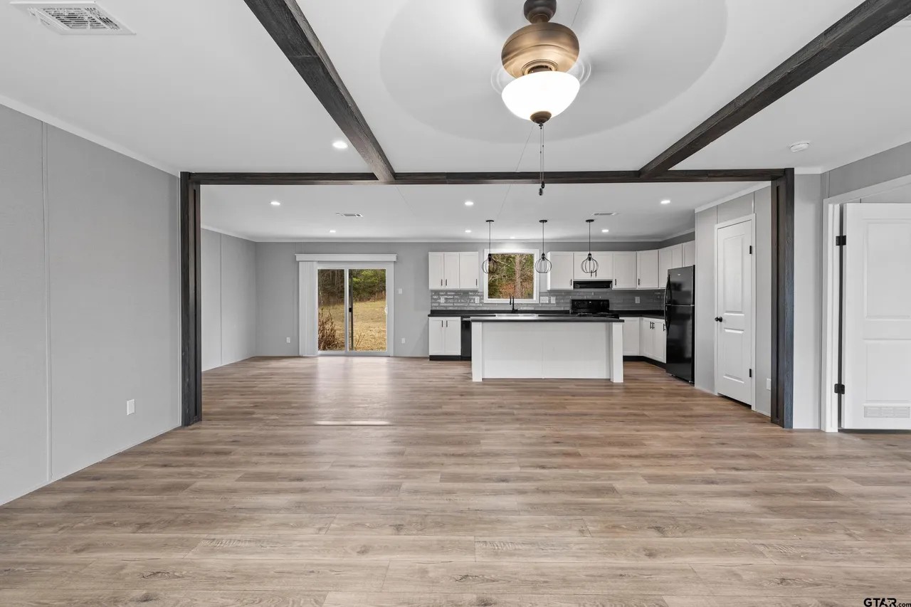 Spacious open-concept room with wooden flooring, gray walls, and ceiling beams. Features a modern kitchen with white cabinets and central island, under a ceiling fan.