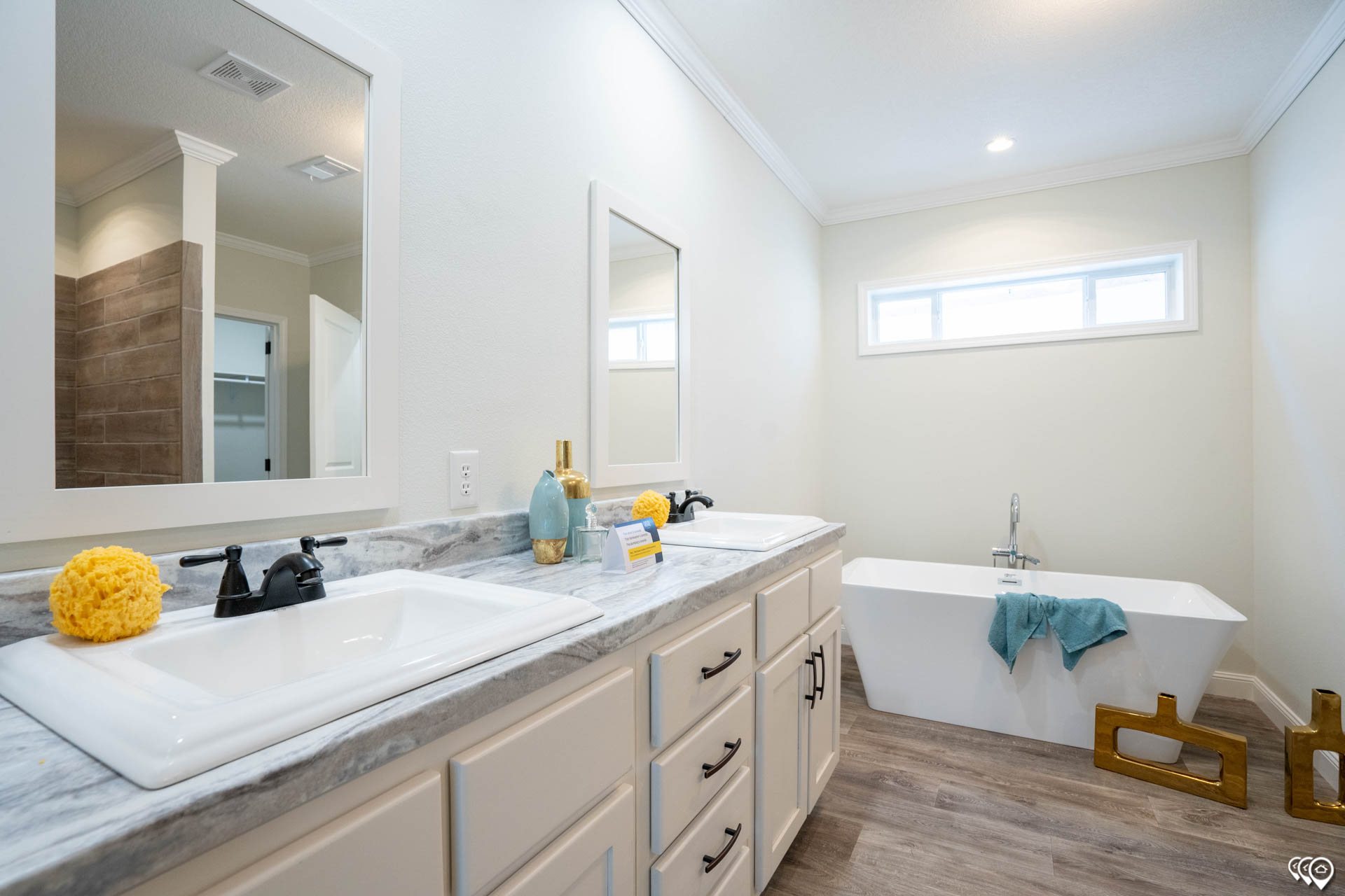 A bright bathroom with a modern feel features a double sink vanity with marble countertop, black faucets, and yellow accents. A white bathtub with a teal towel is set against a light gray wall with a small horizontal window above, creating a calm, inviting atmosphere.