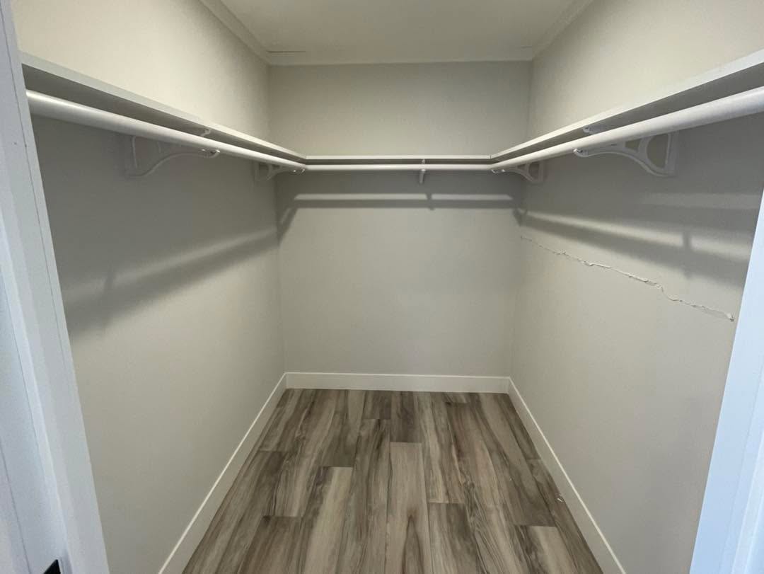 A walk-in closet with white walls, featuring two hanging rods on either side. The floor is wood-paneled, and there's a visible crack on the right wall.