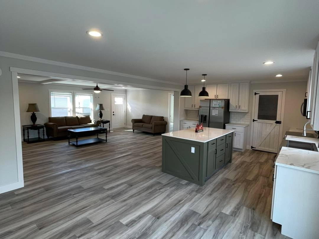 Open-concept kitchen and living area with gray wood flooring. Kitchen features a green island, white cabinets, and pendant lights. Cozy seating in the background.