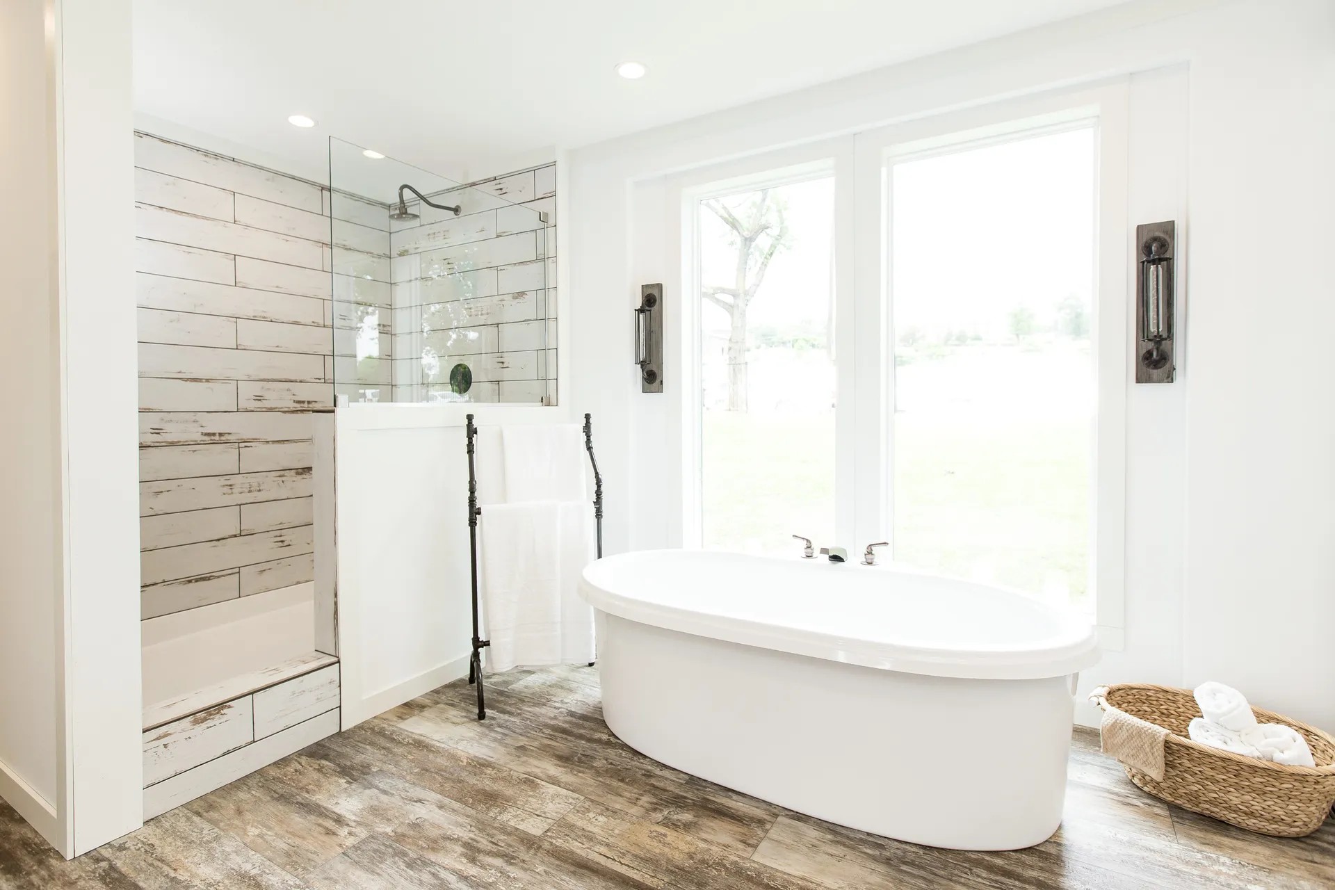 A bright, modern bathroom features a white freestanding tub near large windows, a rustic wood-tiled shower, and a basket with towels, conveying a serene atmosphere.
