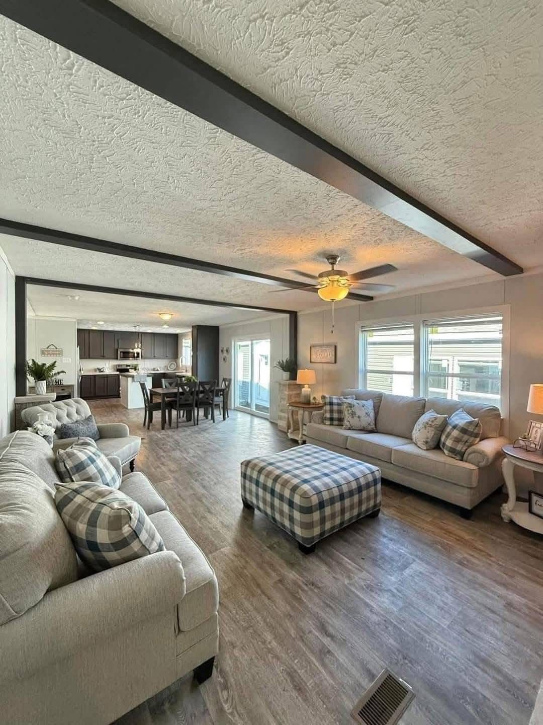 Spacious living room with light gray sofas and plaid cushions, leading to a dining area with a wooden table. Warm lighting and a cozy atmosphere.