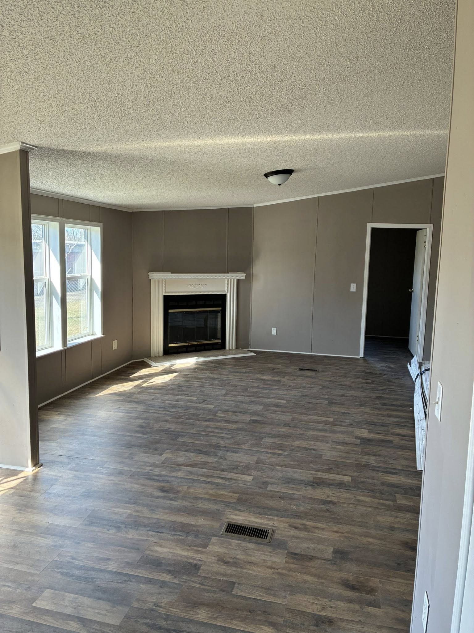 Spacious, empty living room with wooden flooring, beige walls, and a white fireplace. Sunlight streams through three large windows, creating a calm ambiance.