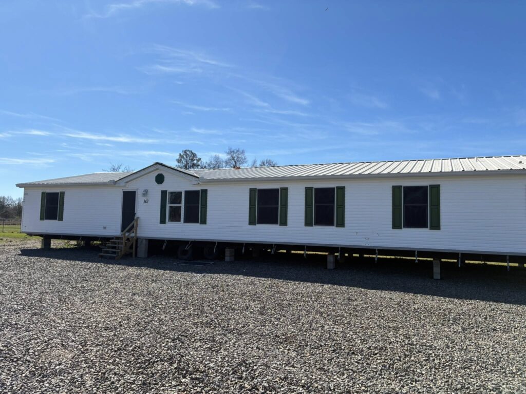 A large white mobile home with a tin roof, green shutters, and small stairs at the front sits on a gravel lot. The sky is clear and blue.