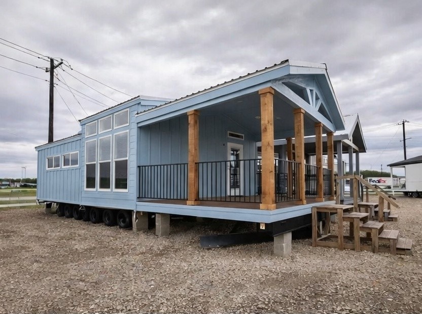 Charming light blue tiny home on wheels with spacious wraparound front porch featuring wooden support columns, black metal railing, covered gable roof with metal roofing, large grid windows for natural light, elevated deck on trailer chassis with sturdy concrete blocks, wooden staircase entry, modern farmhouse exterior design ideal for mobile living, off-grid tiny house living, or affordable housing solution under overcast sky.