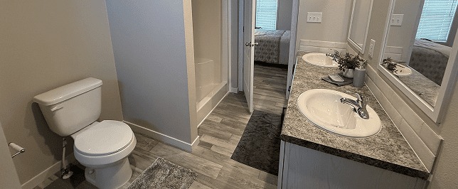 Modern bathroom with dual sinks on a marble countertop, a toilet, and a shower. Neutral tones and a doorway leading to a bedroom convey simplicity.