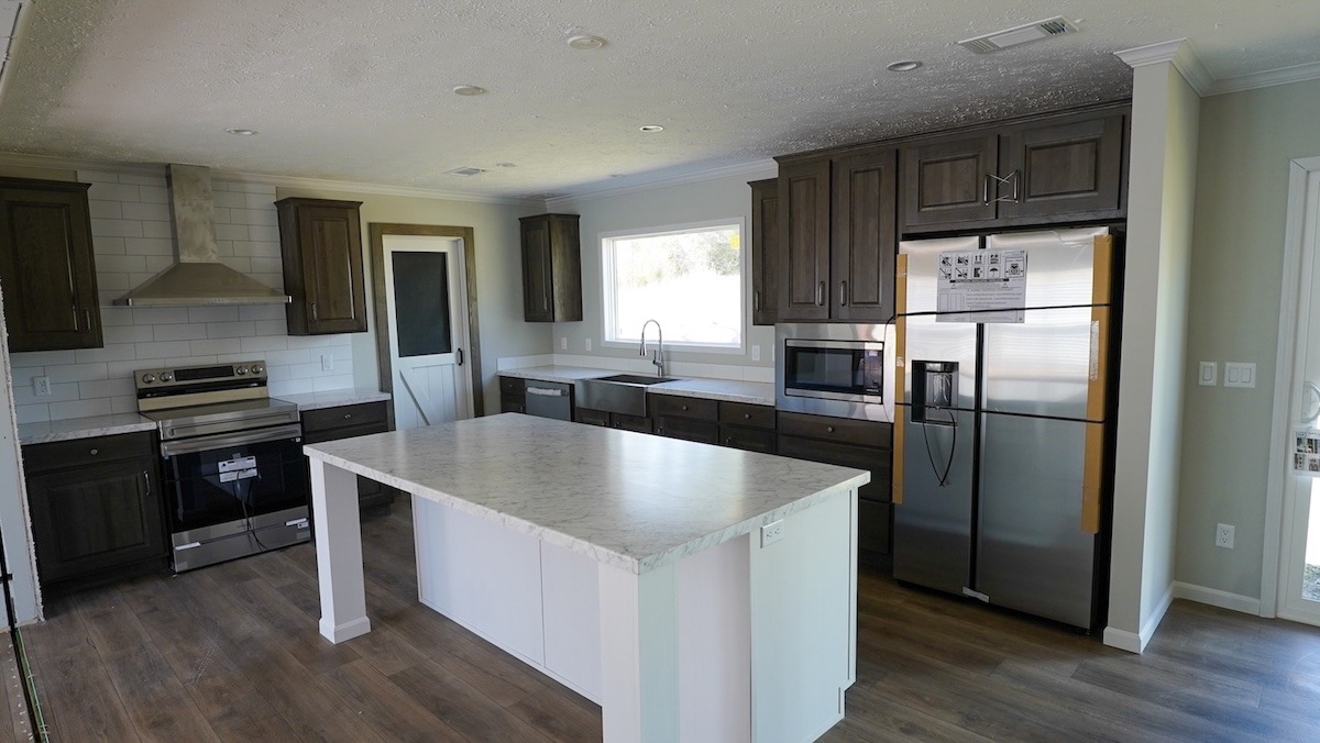 Modern kitchen with dark wood cabinets, stainless steel appliances, and a large white island. Bright window and light wood floors add warmth.