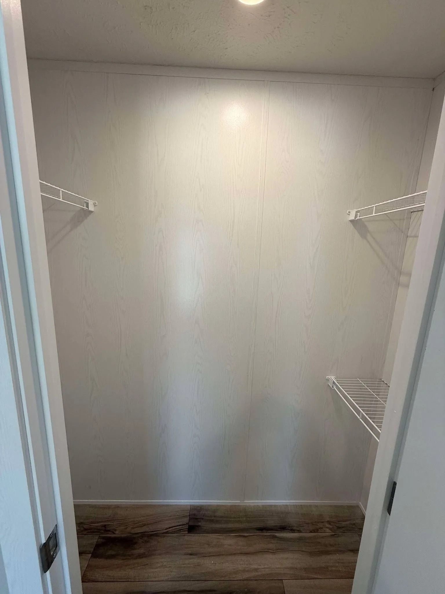 Empty white closet with wire shelves on both sides, wooden floor, and a ceiling light, creating a clean, minimalist look.