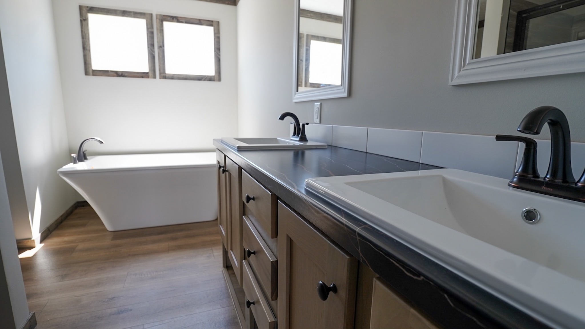Modern bathroom with dual sinks on a black countertop, wooden cabinets, a white freestanding bathtub, and two framed windows, creating a sleek, serene ambiance.