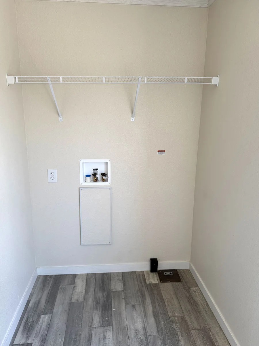 Empty laundry room with gray wood flooring, beige walls, and a white wire shelf above. A small recessed shelf holds containers. A vent is on the right.