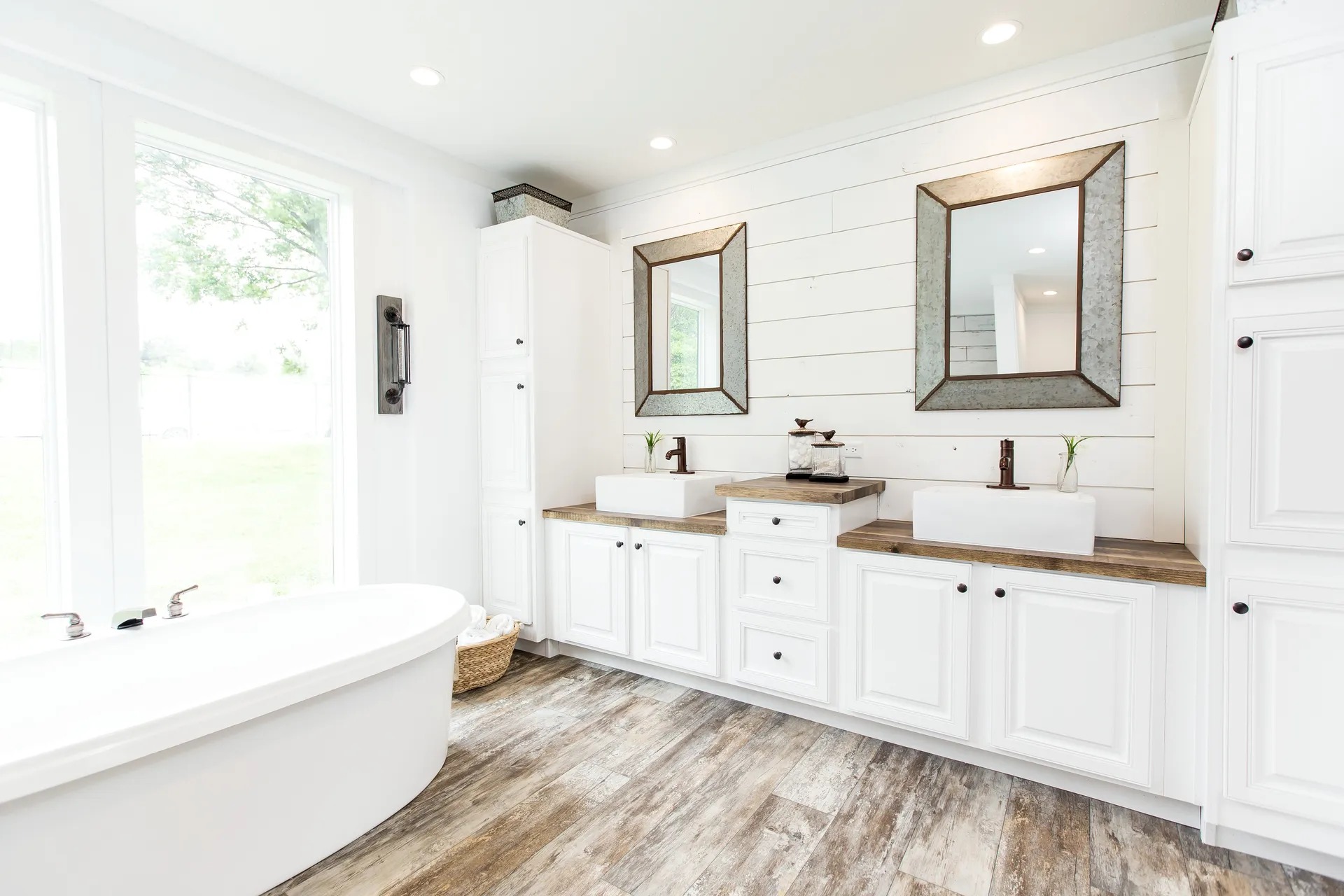 Bright, modern bathroom with shiplap walls, dual sinks with wood countertops, and framed mirrors. Freestanding tub and natural light from large windows.