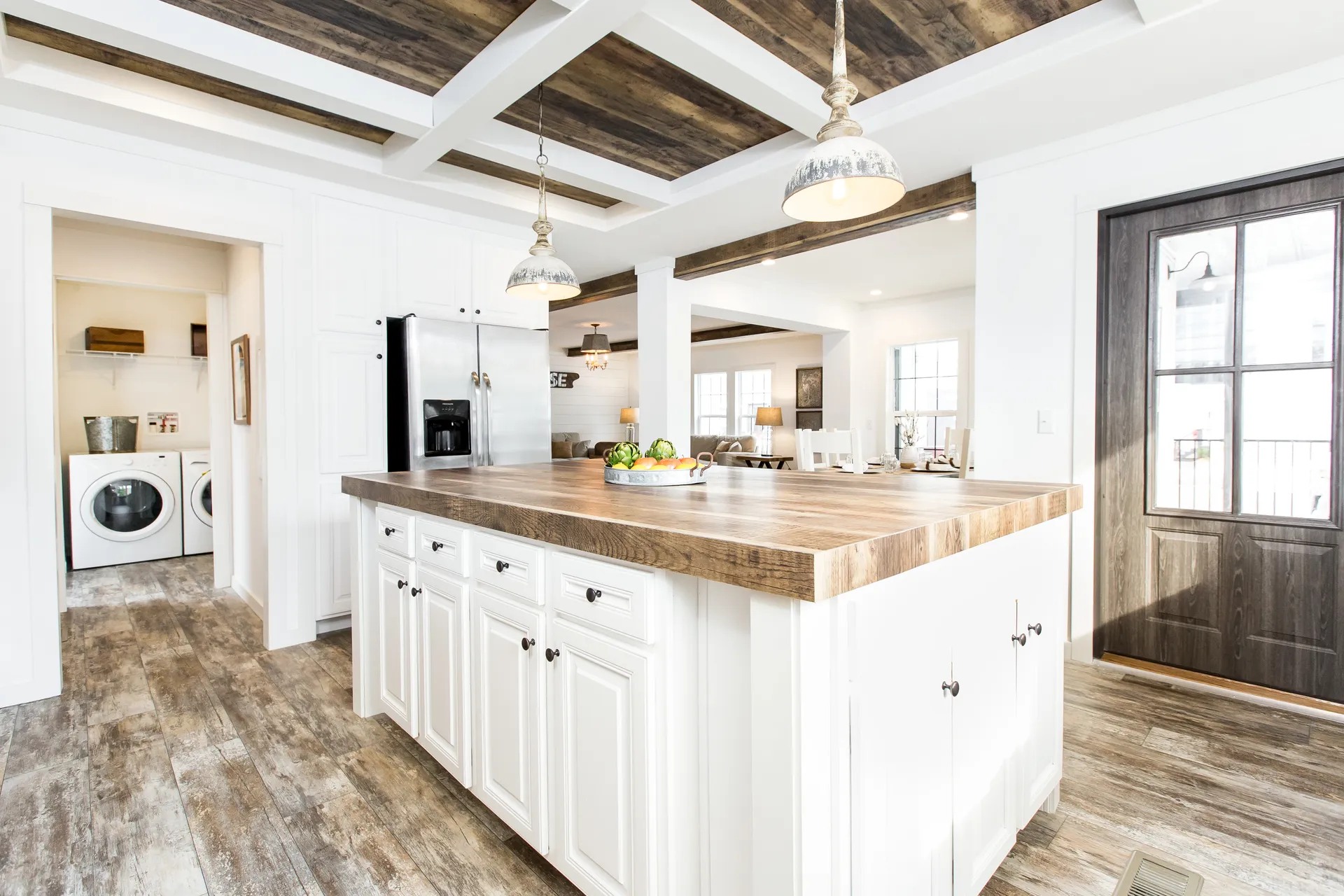 A spacious kitchen with a rustic island featuring a wooden top and white cabinets. Pendant lights hang above. A cozy living area and laundry room are visible.