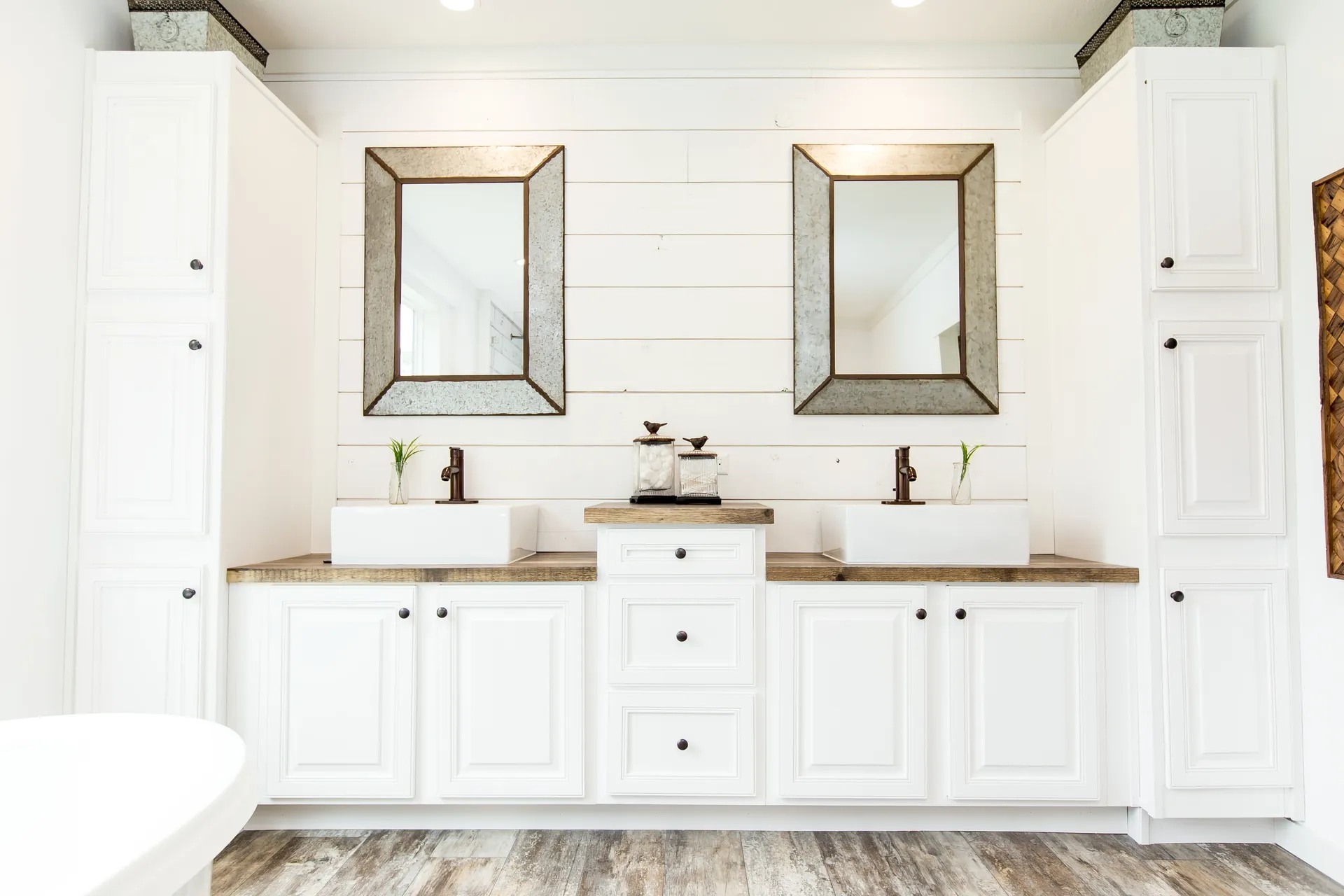 Bright farmhouse-style bathroom with white cabinetry, two wooden-framed mirrors, vessel sinks, brown faucets, and shiplap walls, exuding rustic charm.