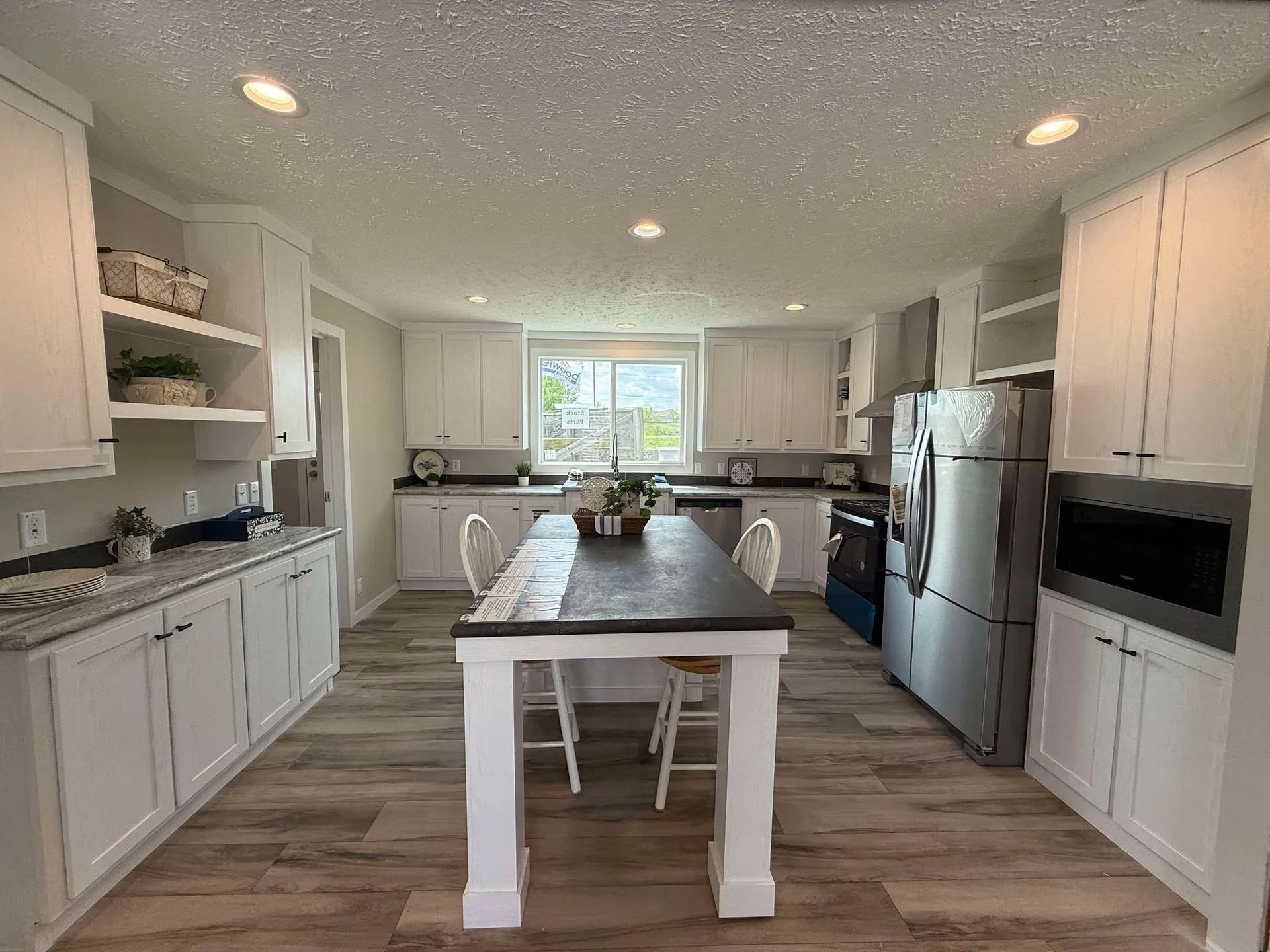 Spacious kitchen with white cabinets, stainless steel appliances, and a central island with seating. Sunlight streams through a large window.