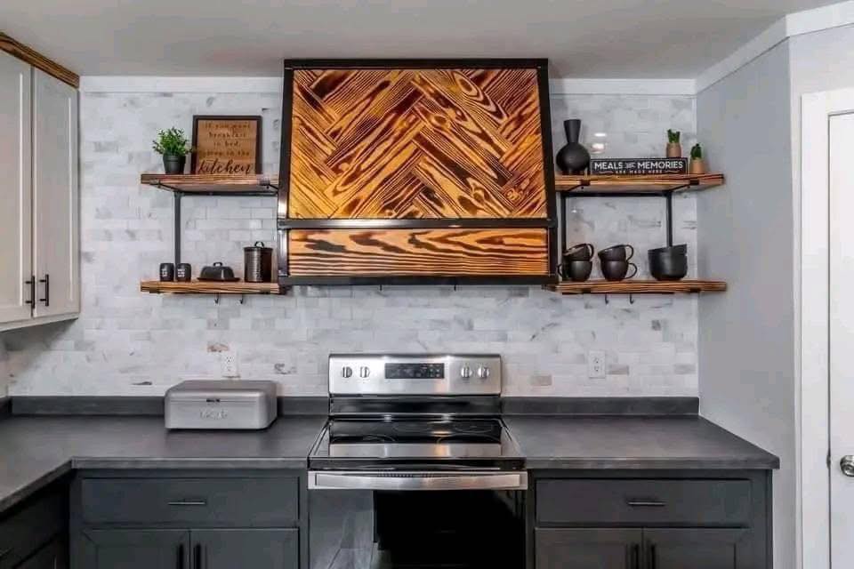 Modern kitchen with gray cabinets, wood herringbone vent hood, stainless steel stove, and open shelves holding plants, books, and black dishes.
