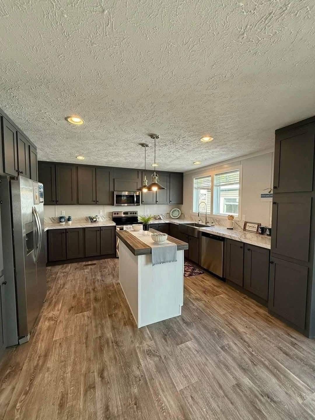 Modern kitchen with dark cabinets and light countertops, featuring a central island with pendant lighting. Stainless steel appliances and wood flooring add a contemporary, cozy feel.