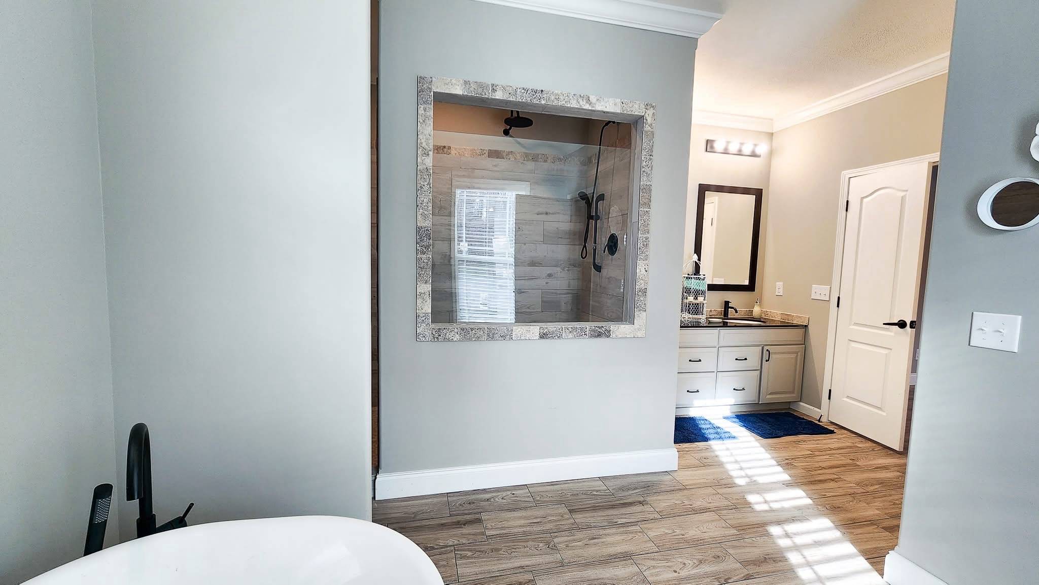 A modern bathroom with light wood flooring. Features include a freestanding white tub, a large walk-in shower with stone accents, and a double vanity with bright lighting, evoking a clean, minimalist feel.