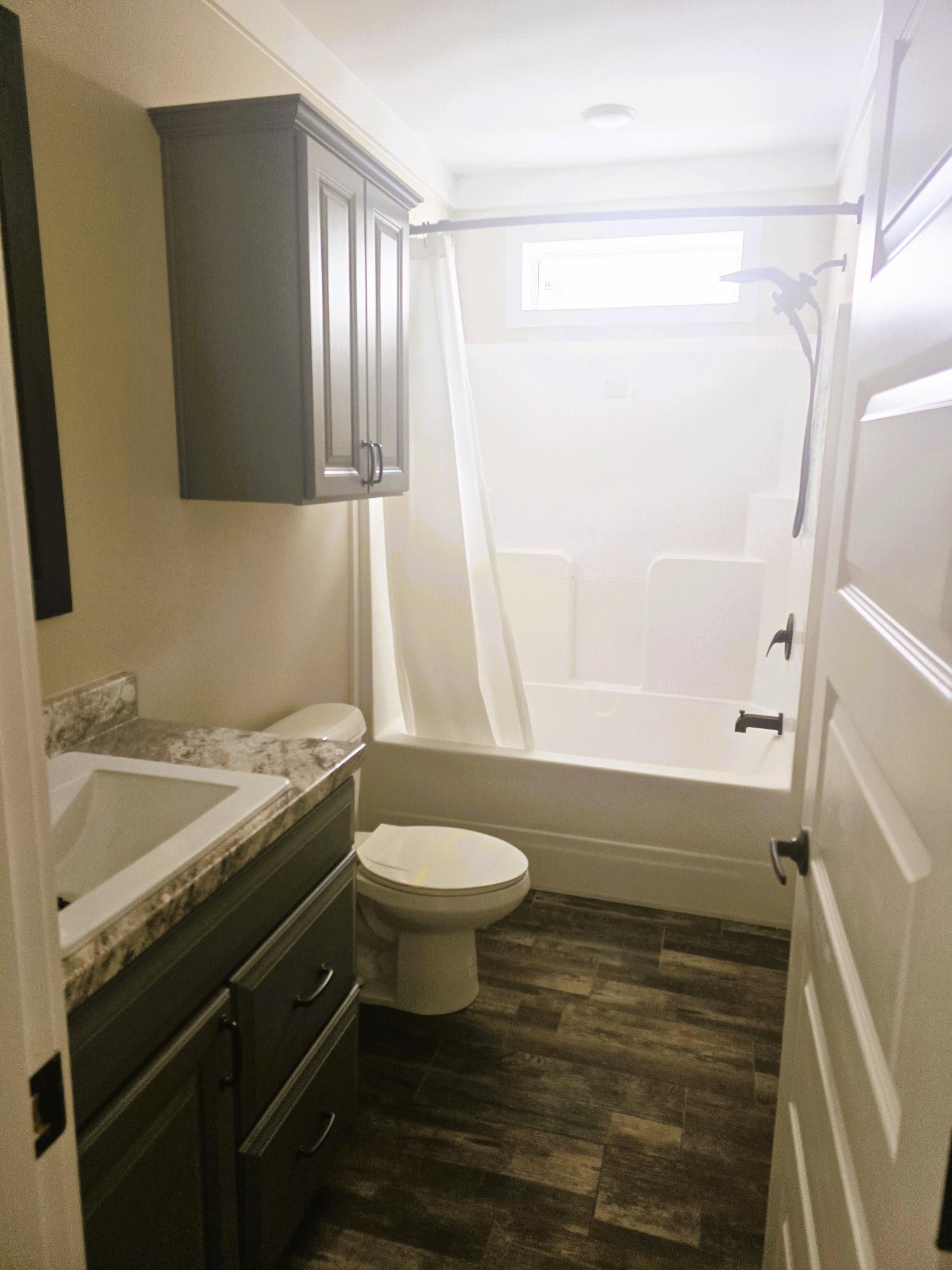 Compact bathroom with a shower-tub combo, white curtain, and a small window. Features dark wood cabinets, a marble countertop, and wood-patterned flooring.
