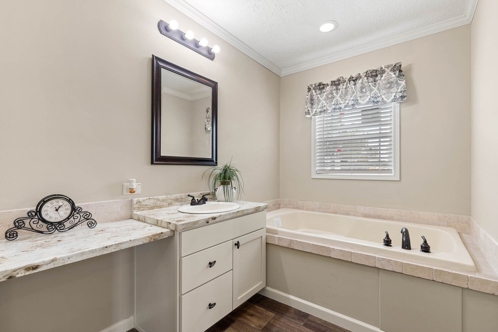 A cozy bathroom features a marble-topped vanity with a mirror, potted plant, and decorative clock. Beside it is a large bathtub with a floral curtain above, soft lighting, and a warm, inviting tone.
