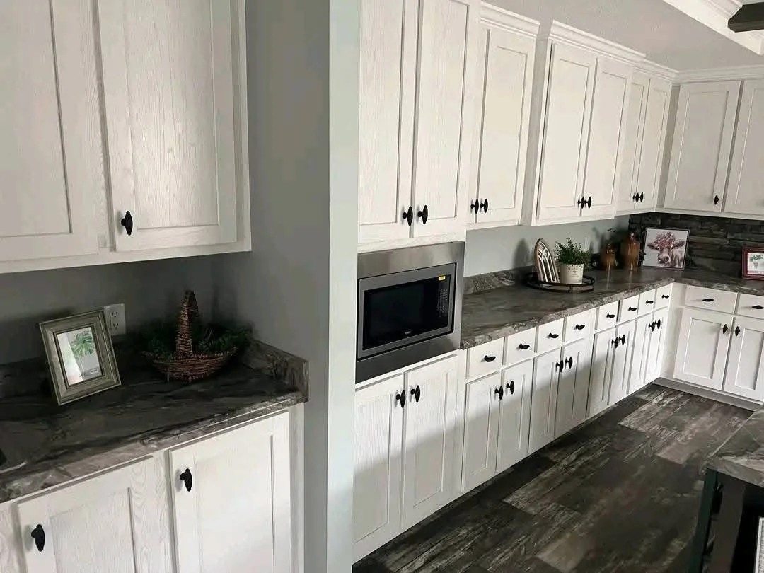 Modern kitchen with white cabinets, gray marble countertops, and a built-in microwave. Decorative plants and framed pictures adorn the surfaces.