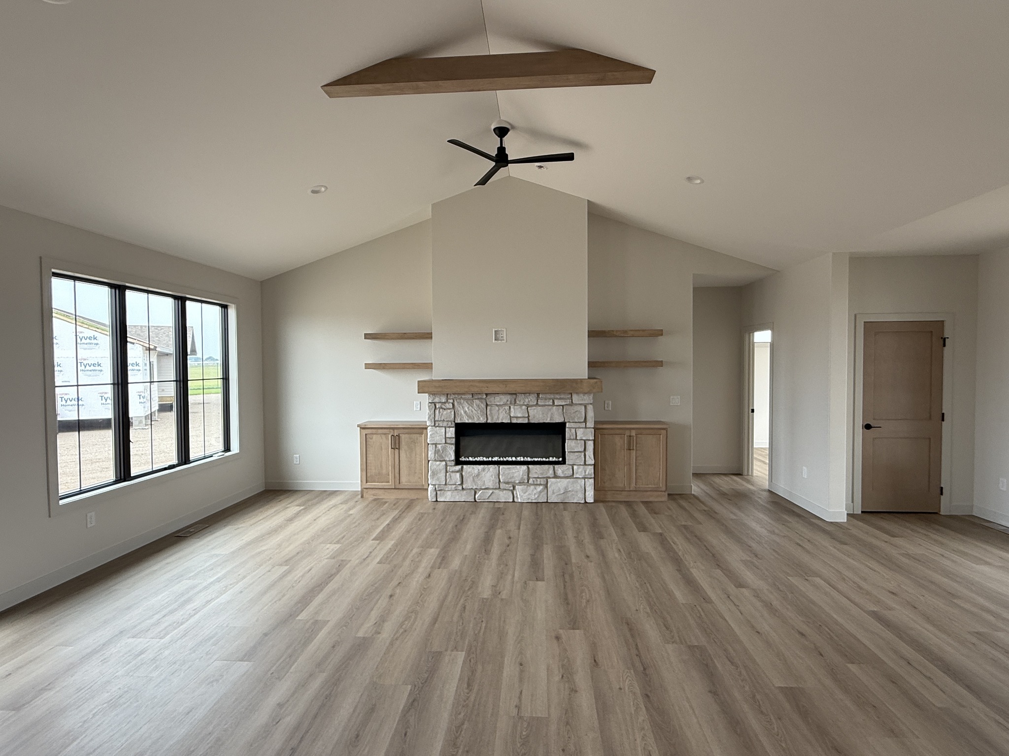 Spacious living room with light wood flooring, a stone fireplace, wooden ceiling beam, and large window. Minimalist shelves and a relaxed ambiance.
