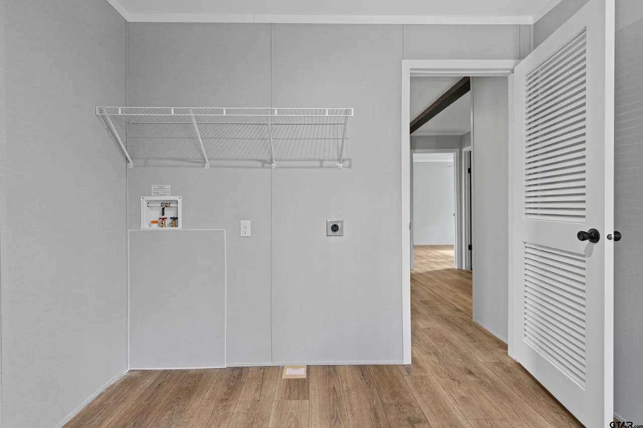 Empty laundry room features light gray walls, a wire shelf, and wood floor. A door on the right opens to a hallway, creating a minimalistic feel.