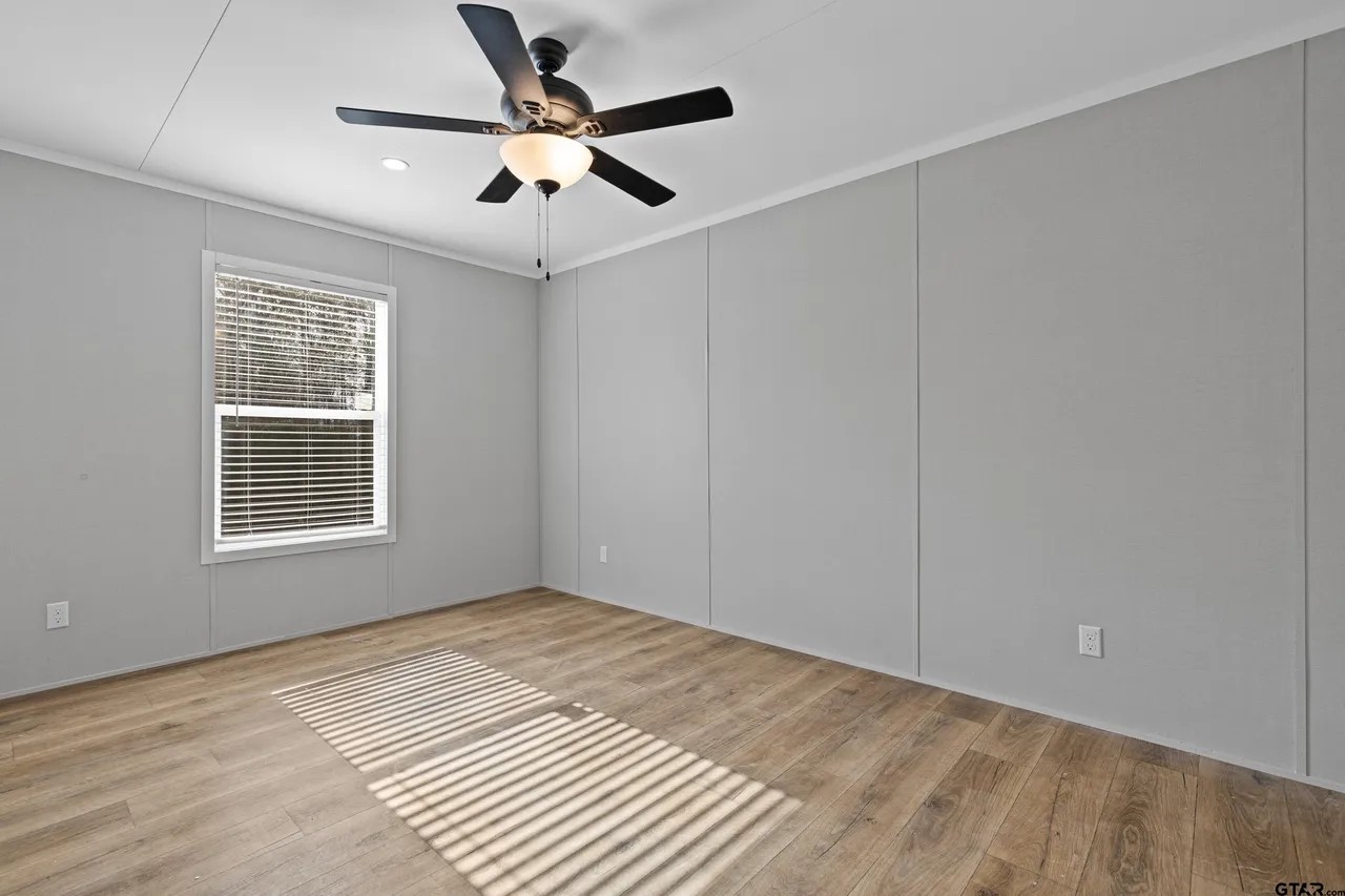 A minimalist, empty room with light gray walls and wood floors. It features a ceiling fan with a light, and sunlight streams through a window, casting shadows.