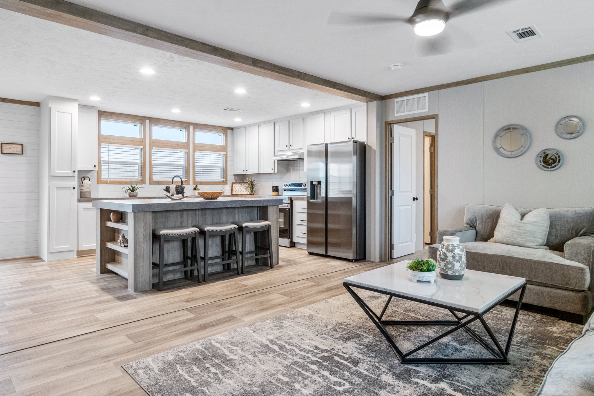 Modern living room and kitchen with a cozy, open layout. Features a gray sofa, sleek coffee table, bar stools, and stainless steel appliances. Bright and inviting atmosphere.