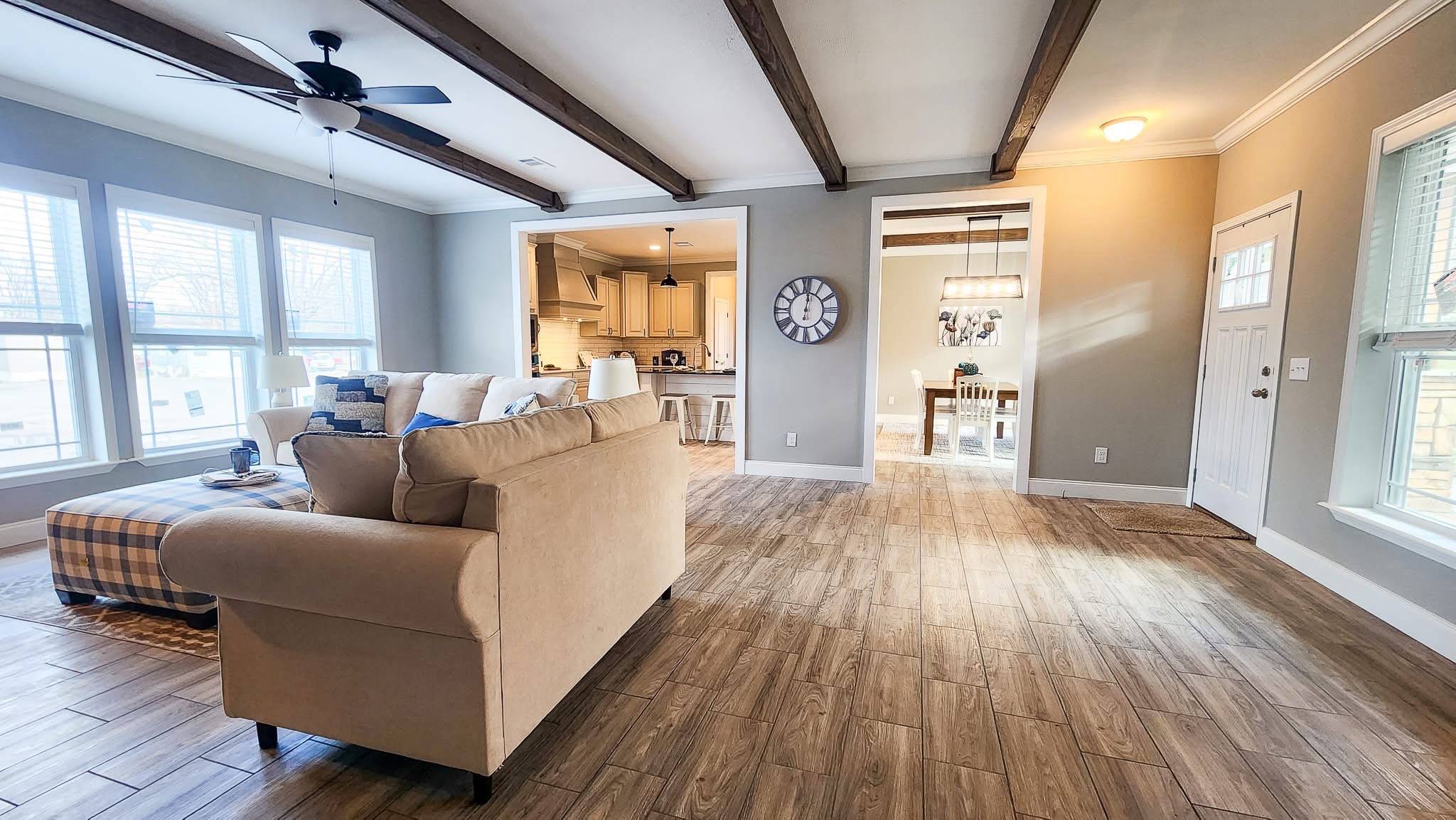 A spacious living area with a beige sofa and plaid ottoman. Wooden ceiling beams and flooring add warmth. An open kitchen and dining room are visible, featuring a large wall clock and ample natural light.