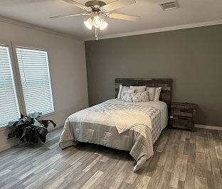 A cozy bedroom with wood flooring features a bed with neutral bedding against a green accent wall. A plant and natural light from a window add warmth.