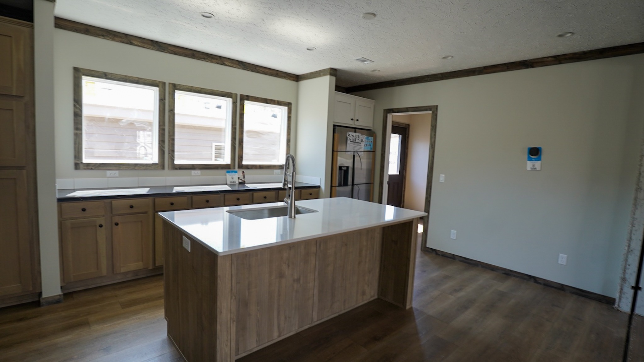 Modern kitchen with a wooden island and sink in the center. Light brown cabinets and stainless steel appliances line the walls. Large windows provide natural light on the wood flooring, creating a bright and welcoming atmosphere.