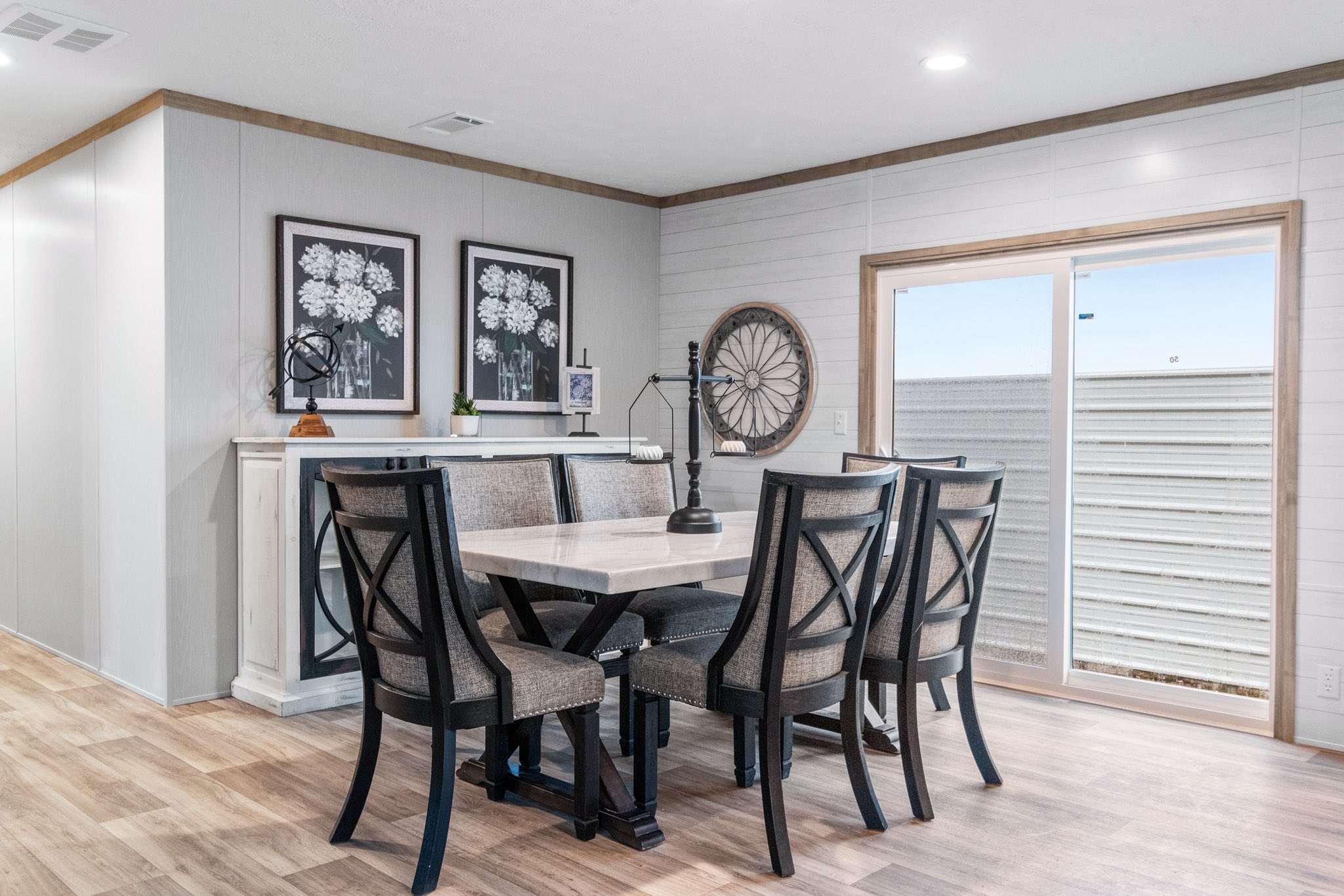 Modern dining room with a rectangular light beige table, surrounded by six cushioned chairs. Two floral framed prints hang on the wall above a console. A large decorative wheel and sliding glass door provide an airy, inviting atmosphere.
