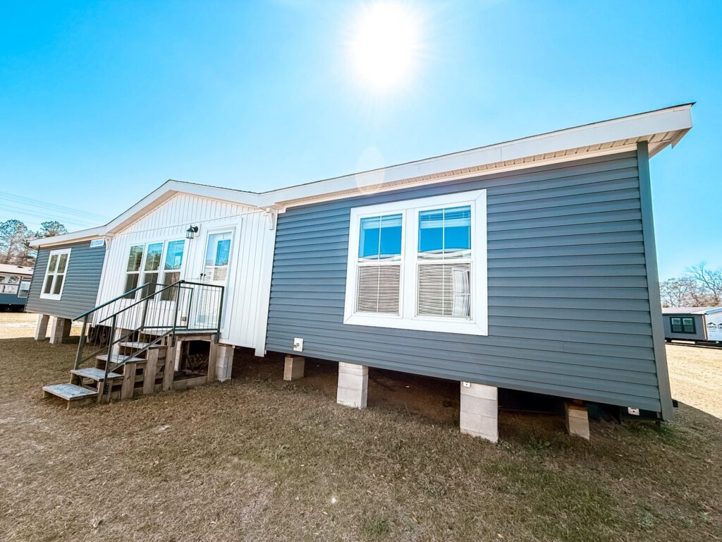 A single-story manufactured home with gray siding and white trim, raised on concrete blocks. Sun shines brightly overhead, casting shadows on a grassy lawn.