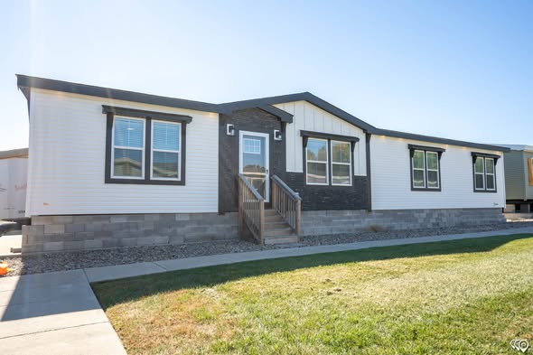 Modern white and gray manufactured home with a stone foundation, large windows, and concrete steps. Sunny day, grass lawn, and a sidewalk in front.