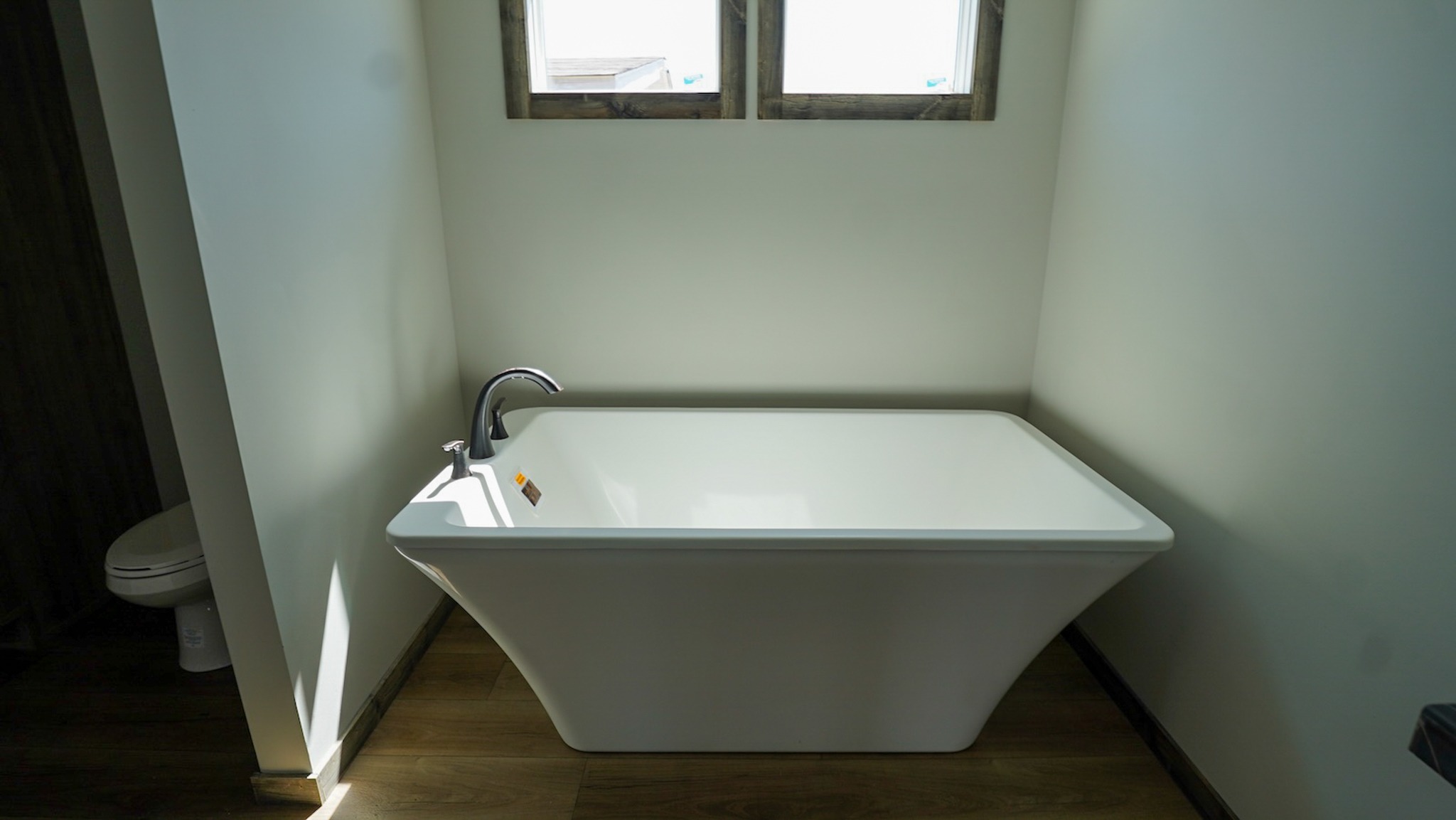 Modern bathroom with a sleek, white freestanding bathtub under two small windows. The room is minimalistic with light walls and wood floors.