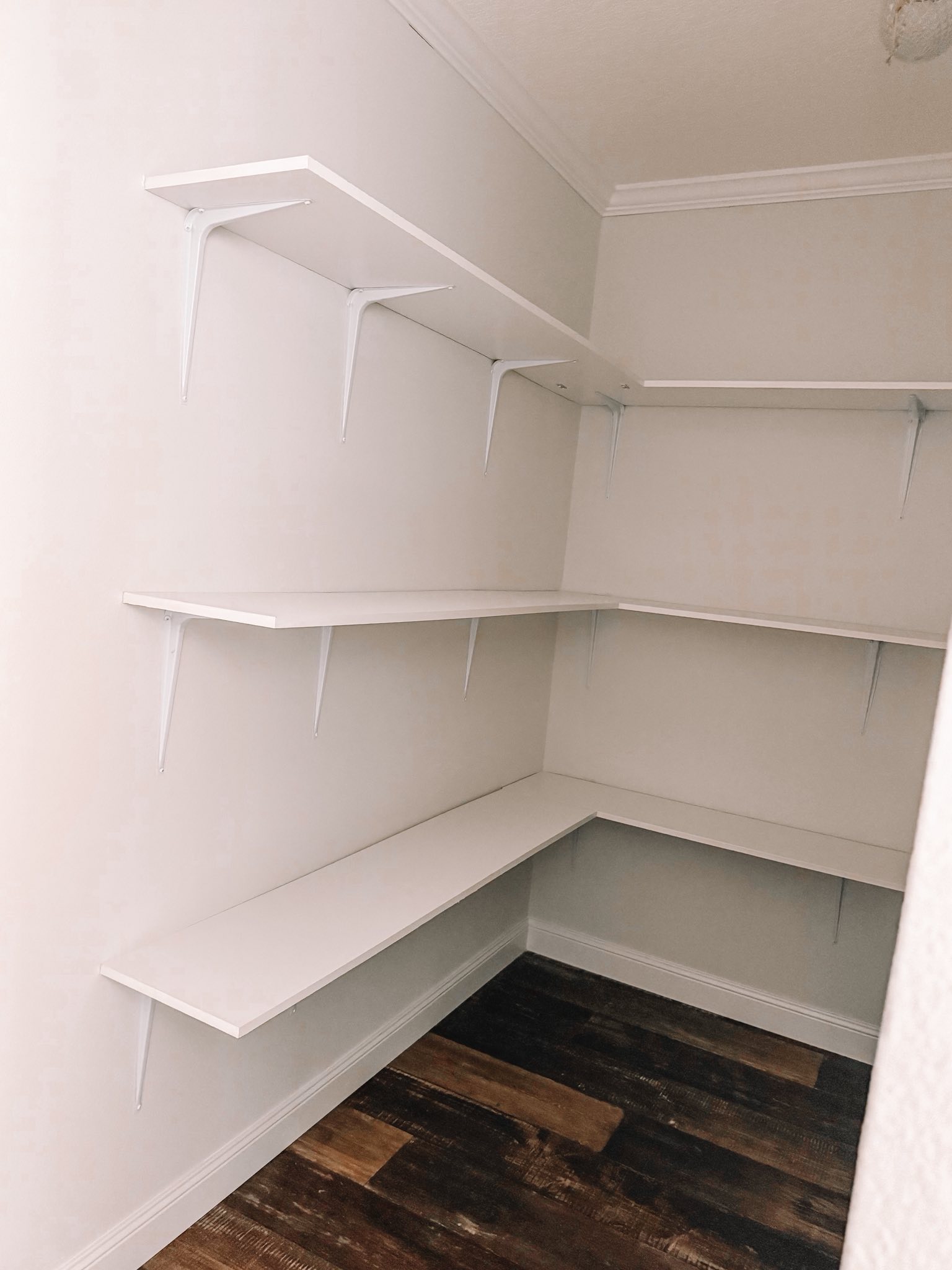 A small pantry with light walls features three tiers of white wooden shelves arranged in an L-shape. The dark wood floor adds contrast to the bright space.
