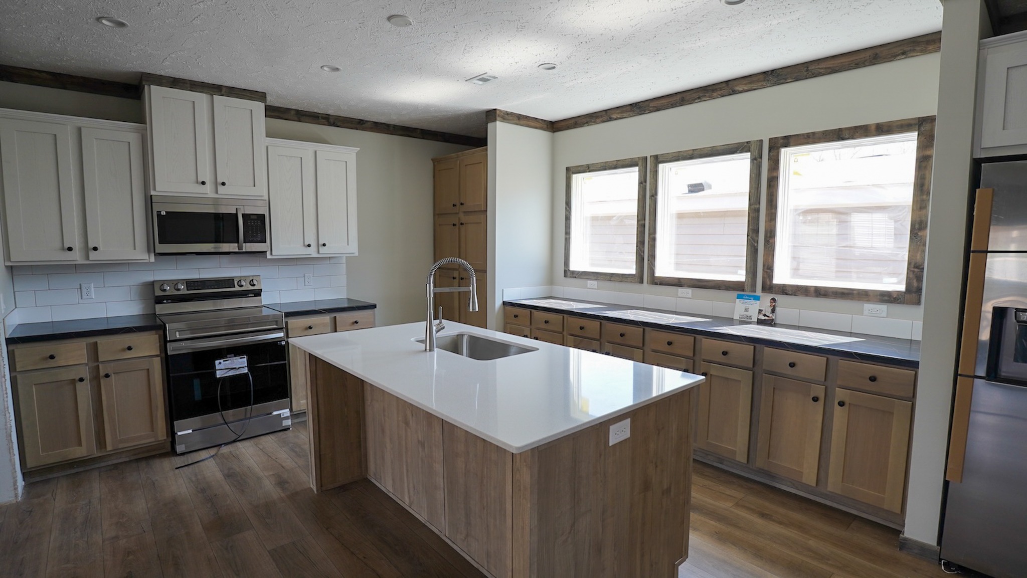 Modern kitchen with wood flooring and white walls. Features include white cabinetry, stainless steel appliances, a central island with a sink, and large windows providing natural light.