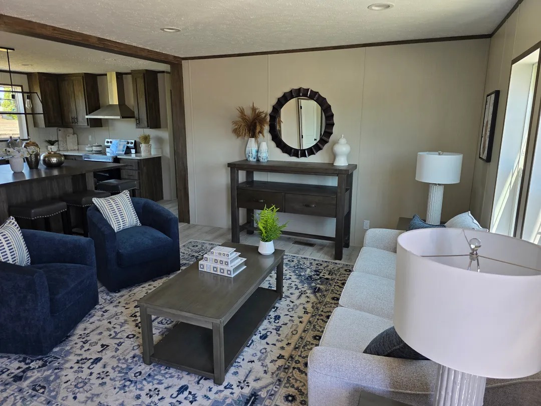 A cozy living room with a light gray sofa, two navy armchairs, and a patterned rug. A round mirror and decor adorn a console table. Warm, inviting ambiance.