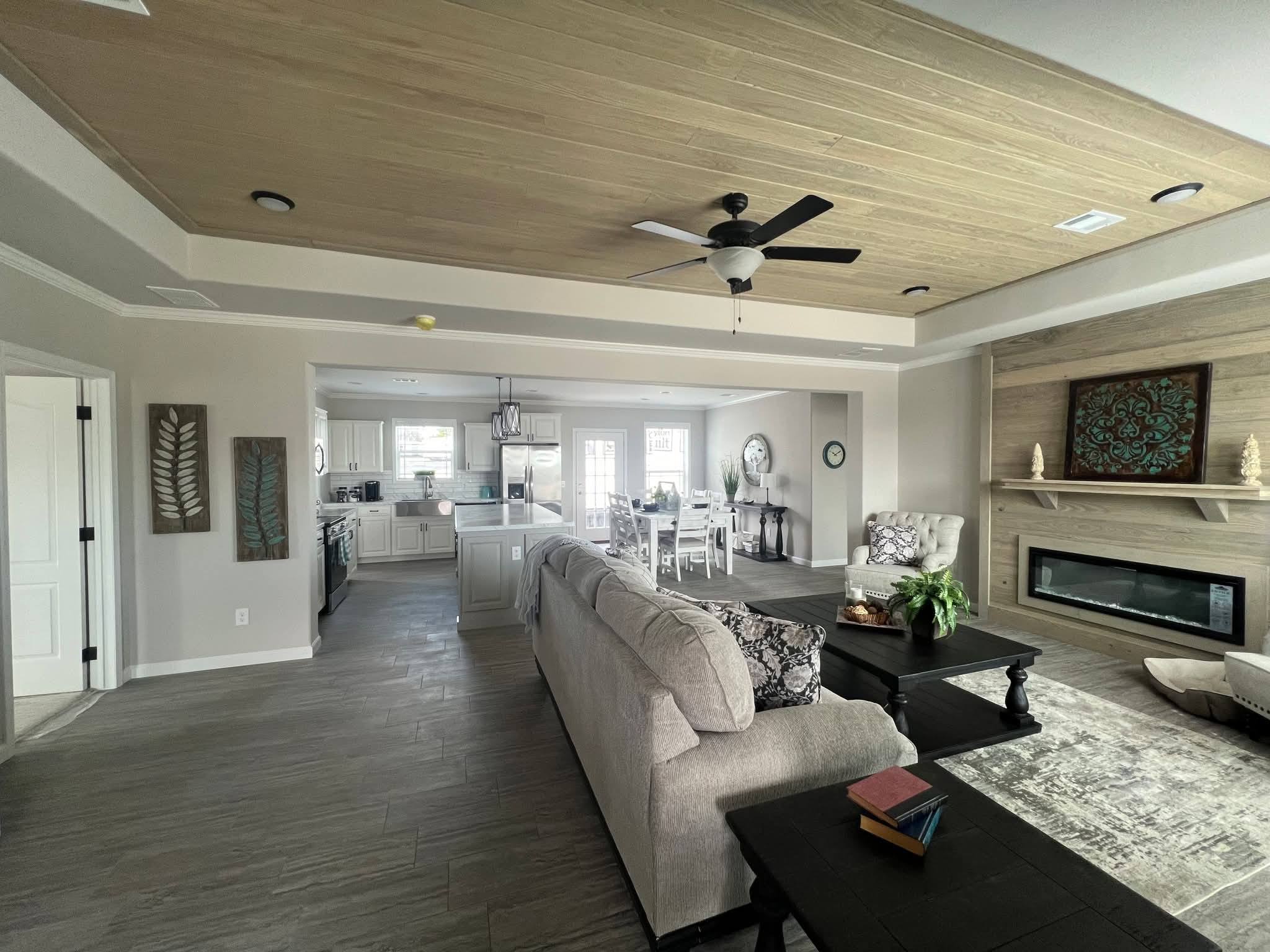 Modern open-plan living room and kitchen with neutral tones. Features include a cozy grey sofa, dark wood coffee table, wall art, ceiling fan, and sleek fireplace. The space is bright and inviting.