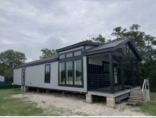 Modern tiny house on raised foundation, featuring large windows and a front porch. The atmosphere is calm with overcast skies and surrounding greenery.
