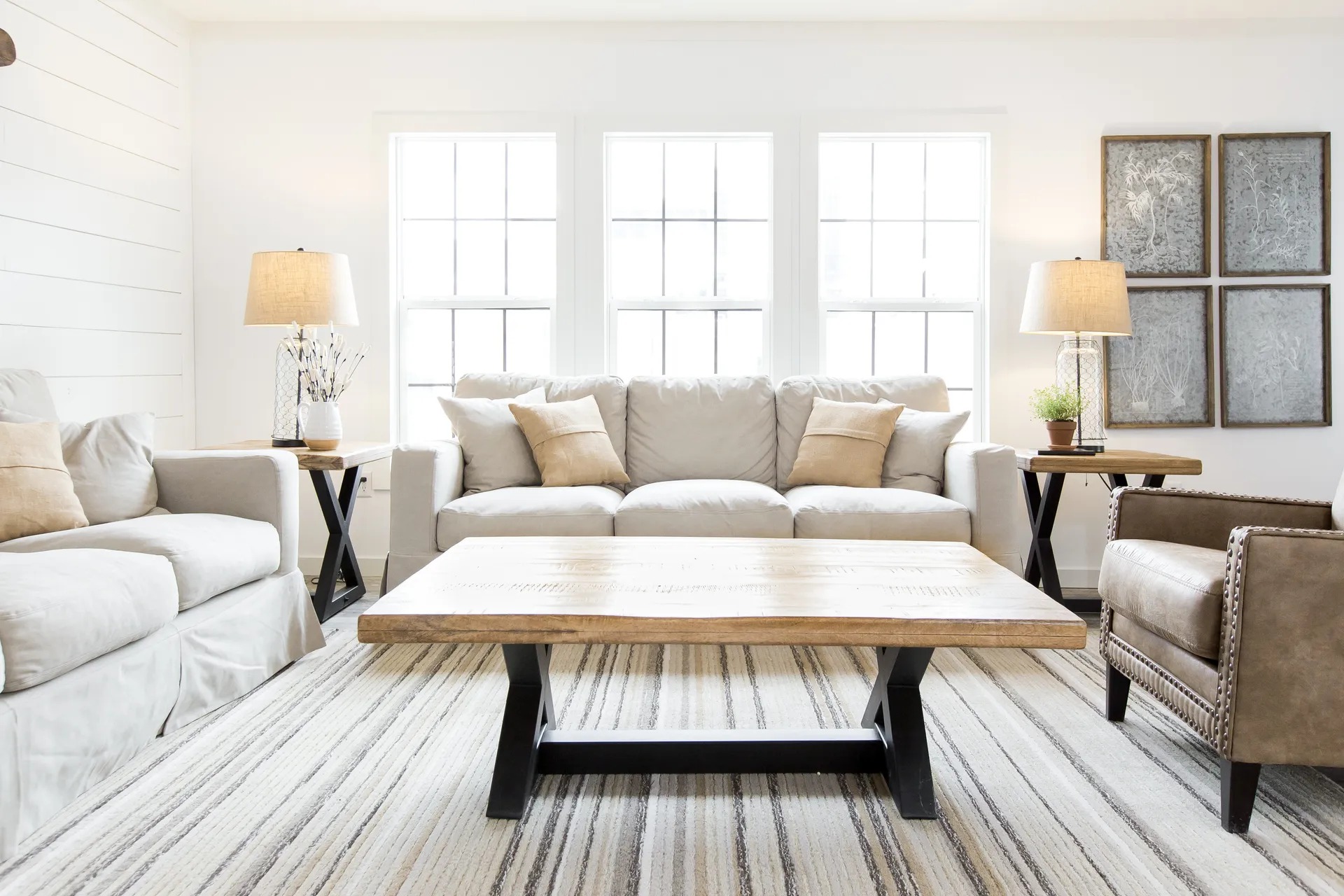 Bright, cozy living room with beige sofas and a brown leather armchair. A wooden coffee table sits on a striped rug, flanked by lamps on side tables. Three large windows light the space.