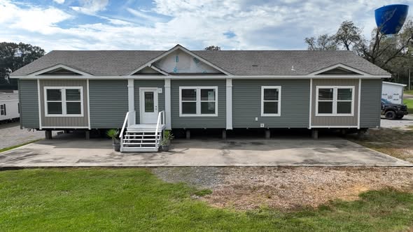 A green modular home with a gray roof and three dormer-like sections sits elevated on a lot with fresh steps leading to the door. A bright blue sky with scattered clouds is overhead.