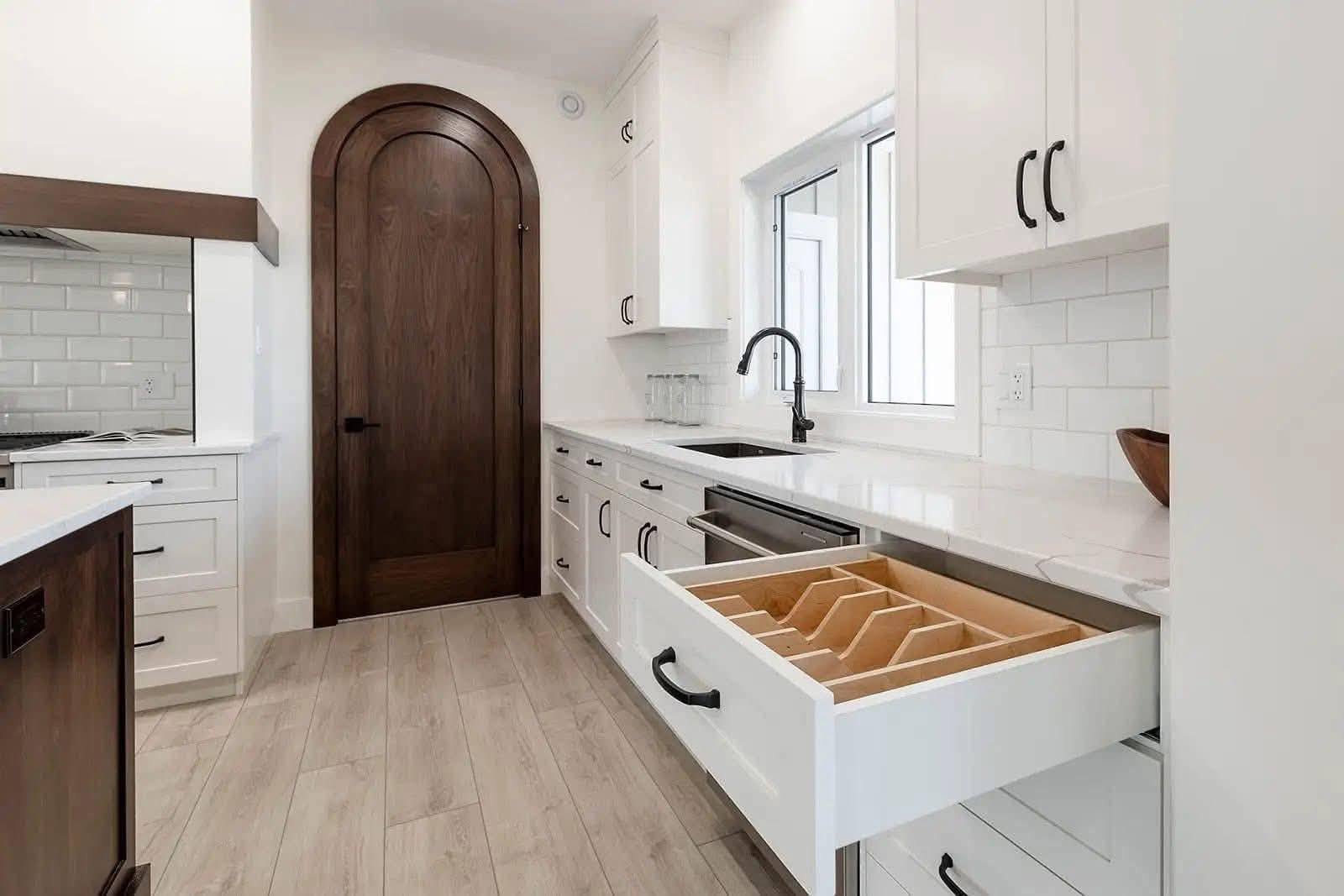 Modern kitchen with white cabinets, open cutlery drawer, and dark wood arched door. Smooth countertops and a stylish black faucet enhance the clean design.