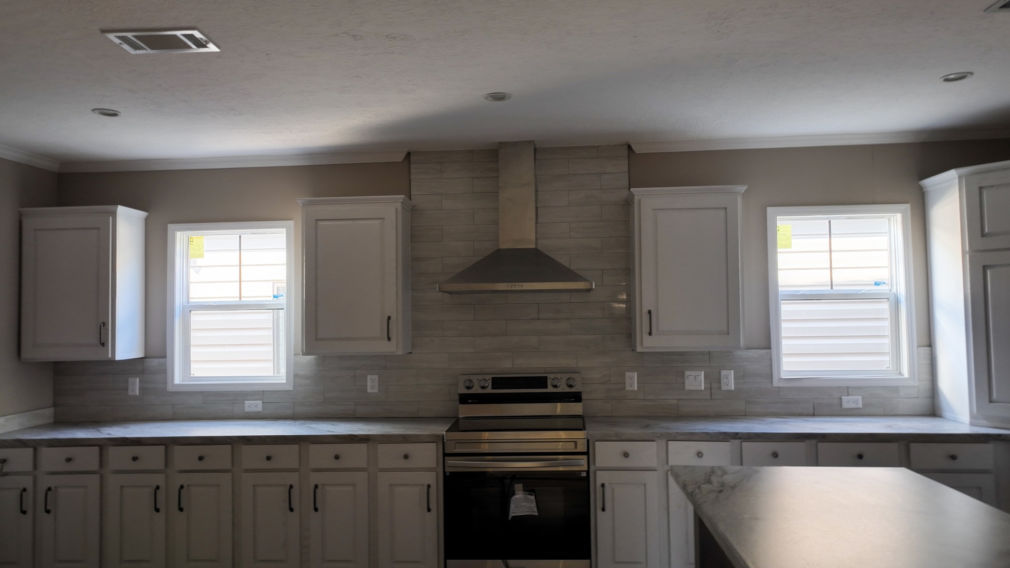 Modern kitchen with gray tile backsplash features stainless steel range and hood. Two windows flank the range, allowing natural light. White cabinets and marble countertops create a clean, serene look.