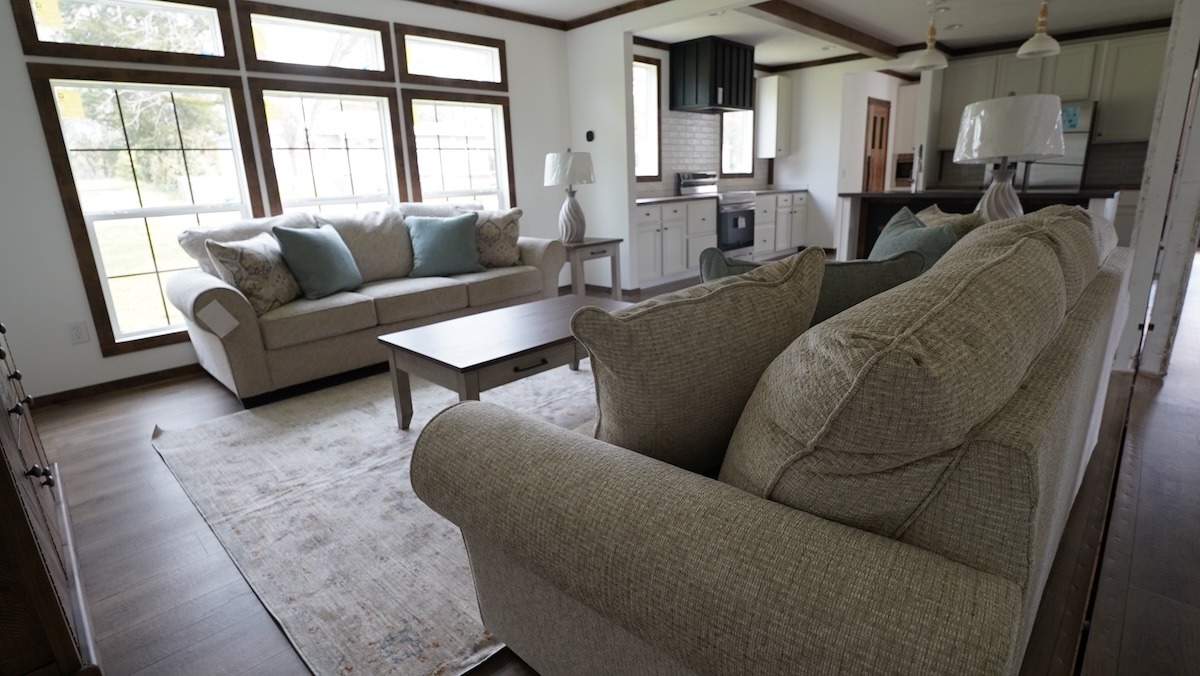 A cozy living room with beige sofas and blue cushions around a wooden coffee table. Large windows fill the space with natural light, enhancing the calm ambiance.