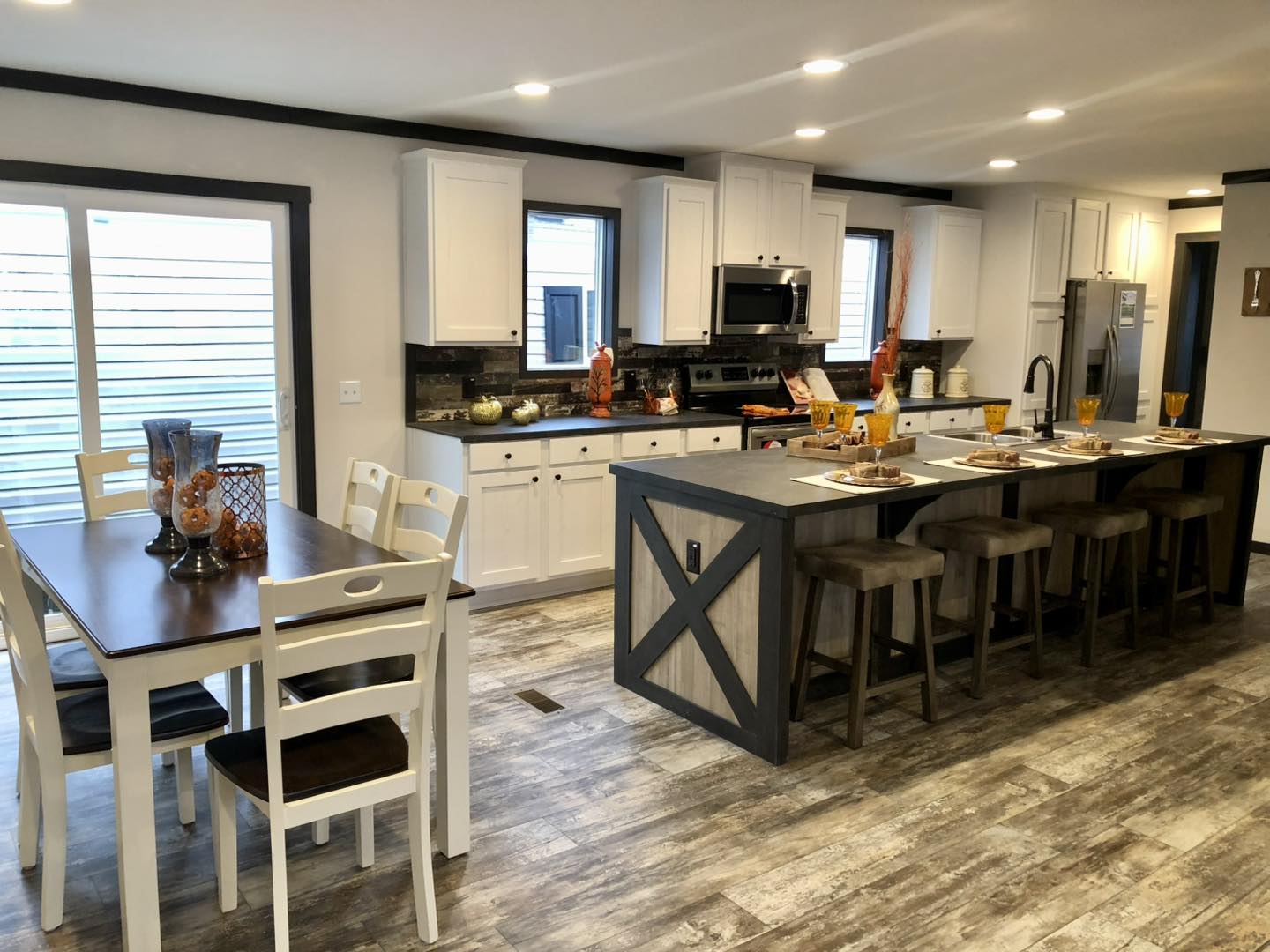 Luxury high-end manufactured home showcase featuring expansive double-wide floor plans, including the white board-and-batten APH 1662C and dark charcoal models with rustic wood shutters. Modern interior highlights include a spacious open-concept living area with an integrated electric fireplace and shiplap accent wall, a gourmet kitchen with a large marble-look island and herringbone tile backsplash, and a custom laundry room with marble-look countertops and cabinetry. The design is elevated by vaulted shiplap ceilings with dark wood beams, a master suite featuring a black accent wall and cowhide rug, a luxury master bathroom with a freestanding soaking tub and LED-mirrored double vanity, and a peaceful covered front porch with a dark wood-plank ceiling and a stylish hanging wicker egg chair.