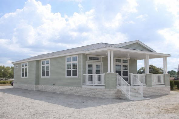 A green, single-story modular home with white trim and a small front porch sits on a gravel lot. The sky is partly cloudy, creating a peaceful ambiance.