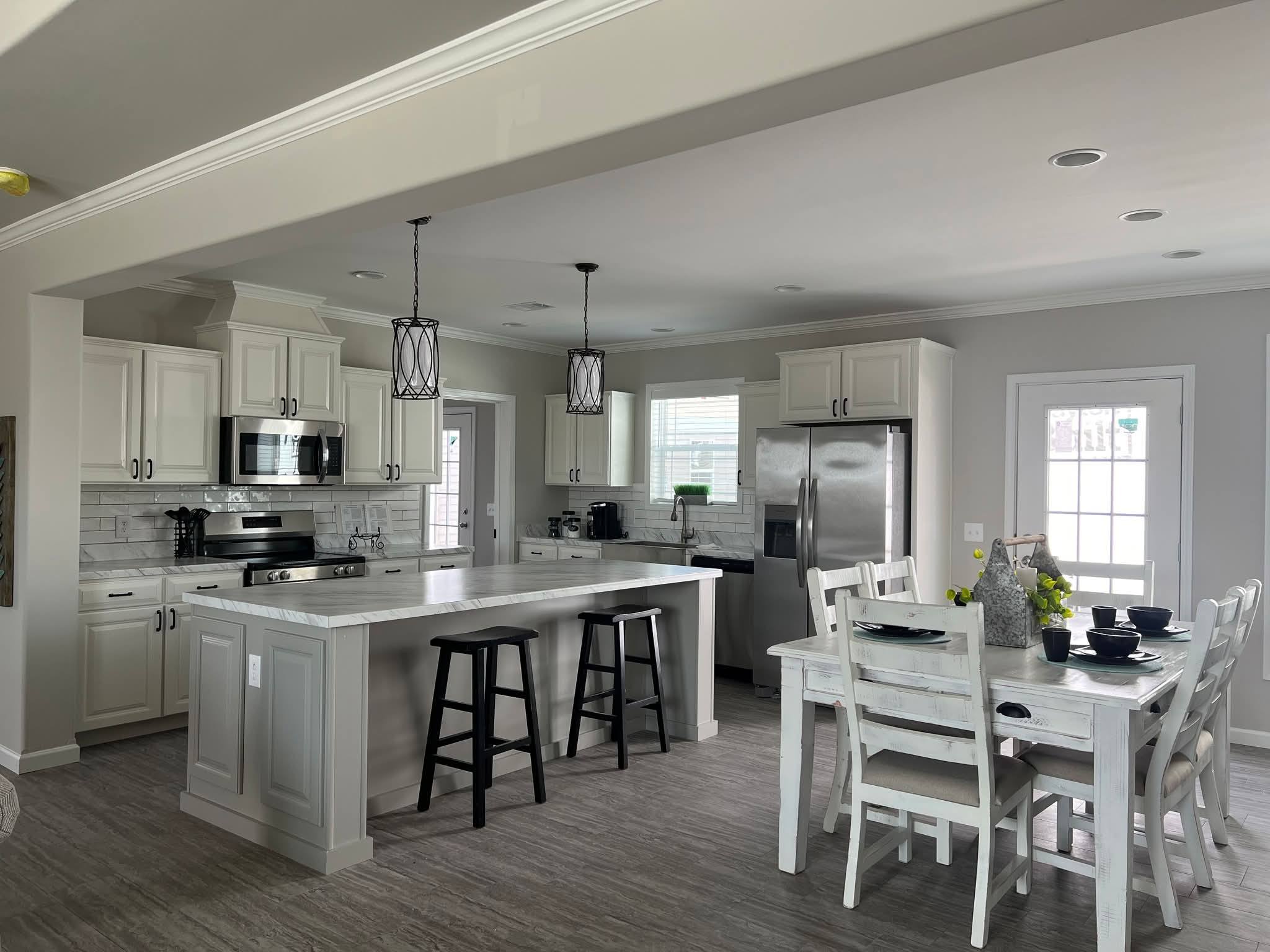 A modern kitchen with white cabinets, stainless steel appliances, and a large island with bar stools. A dining table with black dishware is nearby.