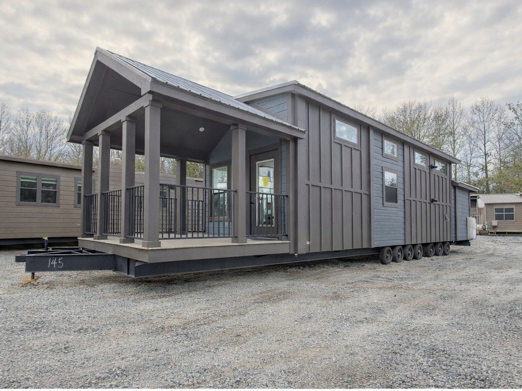 A modern tiny house on wheels features a covered porch with railings and dark wood paneling. It sits on a gravel lot under a cloudy sky, exuding a cozy, inviting vibe.
